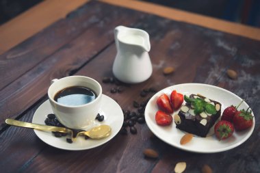 Tahta arka planda beyaz fincanda sıcak kahve, beyaz tabakta taze çilekli Brownie.