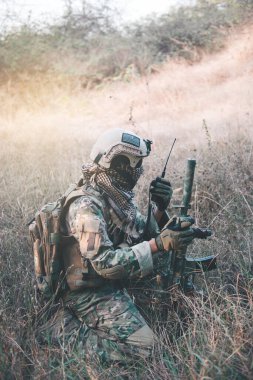 Silahlı bir asker ormanda oturuyordu, telsizle savaş konseptinde destek çağırıyordu.