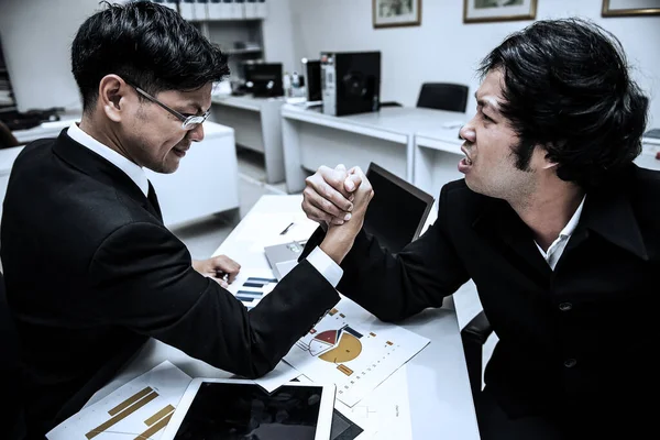 Two asian businessmen arm wrestling together on the table full of sheets of data, laptop and mobile devices,Thailand people