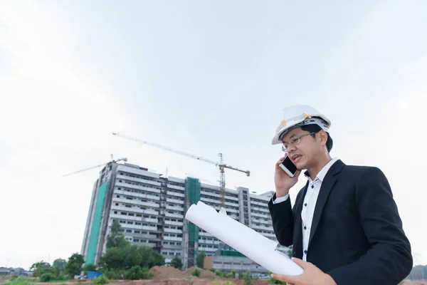 年轻的亚洲工程师在讲电话时 手里拿着大楼的平面图 还有乘客 — 图库照片