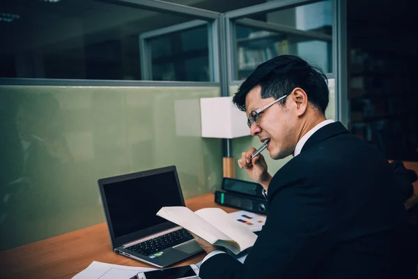 Asian businessman in black suit think idea for work at office,he work overtime alone in a company,dark tone,stress man bite pen