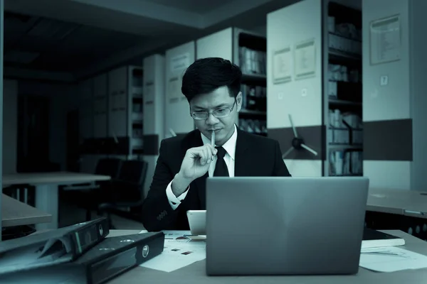 Asian businessman in black suit think idea for work at office,he work overtime alone in a company,dark tone,thailand people