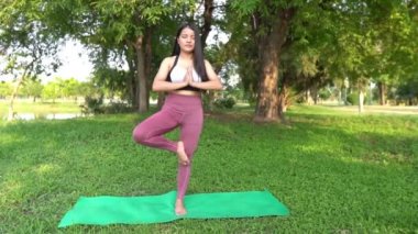 Asyalı genç bir kadın gündüz vakti parkta yoga yapıyor.