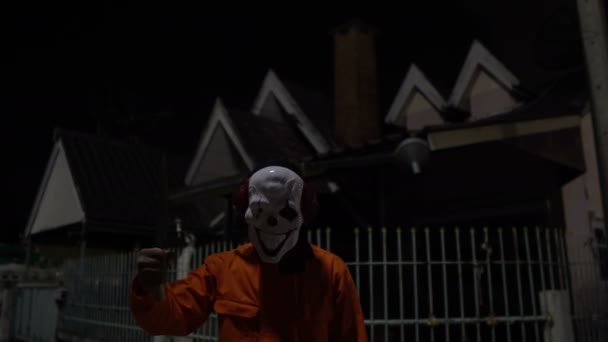 ナイトシーンで武器を持つピエロマスクを身に着けているアジア人男性 ハロウィン祭りのコンセプト オレンジの布で殺人犯のホラー怖い写真 — ストック動画