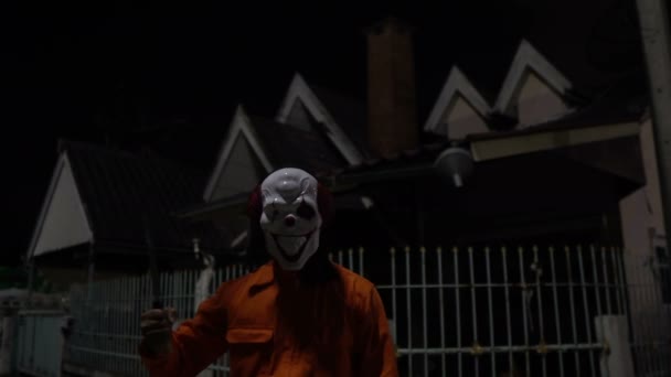 ナイトシーンで武器を持つピエロマスクを身に着けているアジア人男性 ハロウィン祭りのコンセプト オレンジの布で殺人犯のホラー怖い写真 — ストック動画