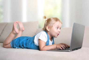 Curiouse çocuk dizüstü bilgisayarı izliyor. Küçük kız not defterine bakarken şaşırdı. Çocuk kanepe kullanım interneti kullanıyor. Teknoloji ya da bilgisayara bağımlılık..