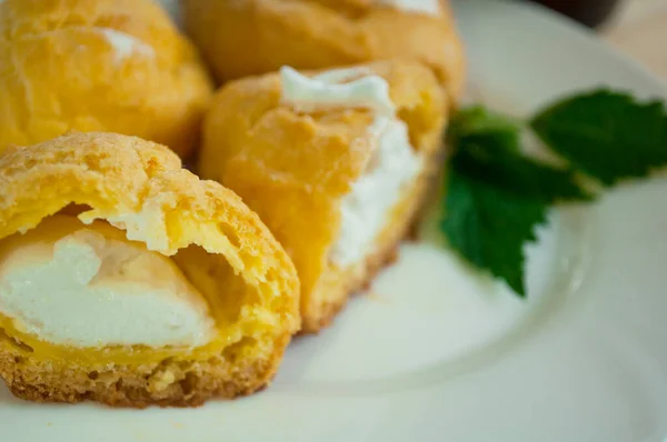 Ahşap arka planda çırpılmış kremayla dolu eklerlerin bir kısmı. Muhallebi kurabiyeleri yakın. Beyaz tabakta pasta, diyet değil.