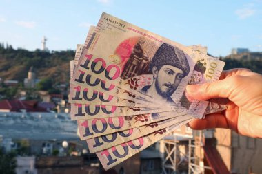 Obverse Side 'da 100 Gürcistan Lari banknotundan oluşan Hand Holding Bunch ve Arkaplanda Bulanık Tiflis Hava Görüntüsü