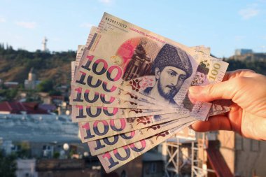 Hand Holding Bunch of 100 Georgian Lari banknotes in Obverse Side with Blurry Tbilisi Aerial View in the Backdrop clipart