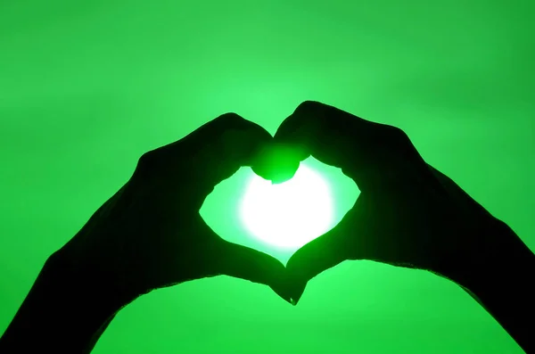 Pop art style silhouette of female's hand posing HEART sign against shiny sun on green colored sky