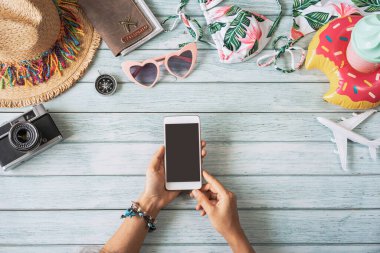 Elinde boş ekran akıllı telefonu, seyahat eden yaz aksesuarları ve ahşap arka planda fotokopi alanı ve seyahat planlama konsepti olan eşyalar tutan genç bir kadın.