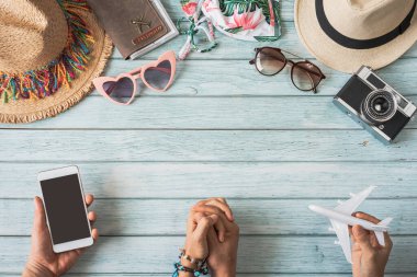Seyahat eden, yaz aksesuarları ve ahşap arka planda fotokopi alanı ve seyahat planlama konsepti olan boş ekran akıllı telefonu olan genç bir çift.