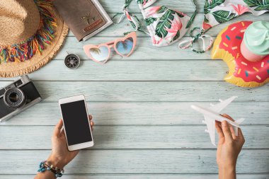 Elinde boş ekran akıllı telefonu, seyahat eden yaz aksesuarları ve ahşap arka planda fotokopi alanı ve seyahat planlama konsepti olan eşyalar tutan genç bir kadın.