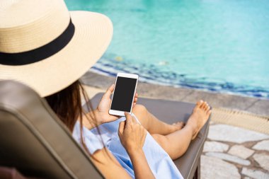 Yaz tatilinin tadını çıkaran genç bir kadın gezgin plaj yakınlarındaki tropik bir tatil beldesinde yüzme havuzunda akıllı telefon kullanarak seyahat yaşam tarzı kavramını kullanıyor.