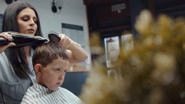 Un drôle de garçon a visité le salon de coiffure. Belle coiffeuse arménienne fait une coupe de cheveux. 4K — Video