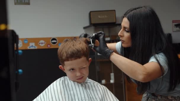 Un drôle de garçon a visité le salon de coiffure. Belle coiffeuse arménienne fait une coupe de cheveux. 4K — Video