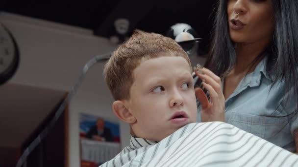 Un drôle de garçon a visité le salon de coiffure. Belle coiffeuse arménienne fait une coupe de cheveux. 4K — Video