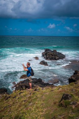 Uçurumun kenarında duran olgun bir adam kayalık kıyıya karşı dalgalı denizin fotoğraflarını çeker.