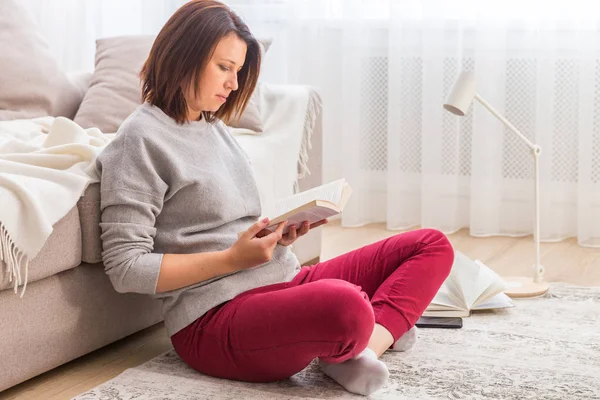 Молода жінка відпочиває на підлозі вдома читаючи книгу — стокове фото