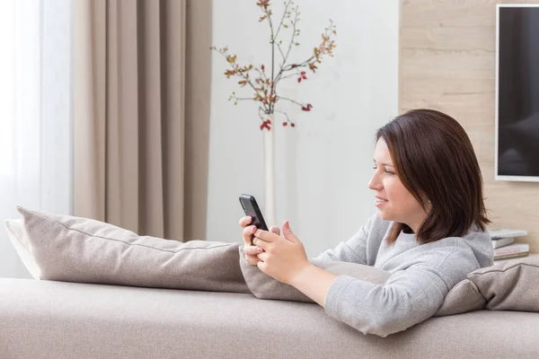 Junge Frau blickt auf ihr Smartphone und lächelt, während sie auf der Couch sitzt — Stockfoto