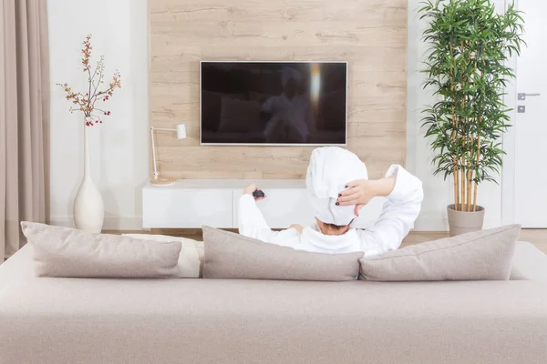 Žena sedí na gauči s ručníkem na hlavě, sledování televize — Stock fotografie