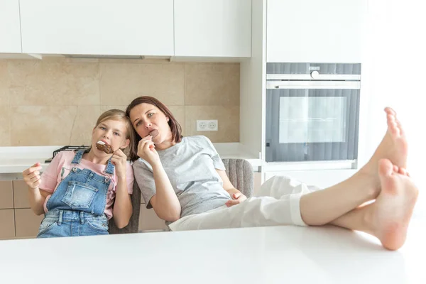 Schattig meisje en haar moeder glimlachen terwijl het eten van ijs in de keuken met benen op een tafel — Stockfoto
