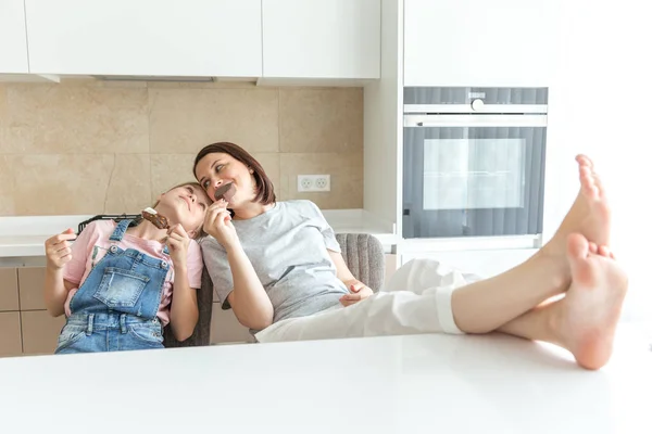 Menina bonito e sua mãe estão sorrindo enquanto come sorvete na cozinha com as pernas em uma mesa — Fotografia de Stock