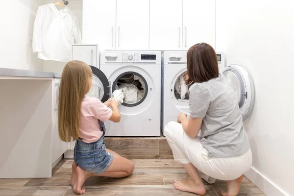 Madre e hija ayudante en la sala de lavandería cerca de la lavadora y secadora quitando la ropa limpia — Foto de Stock