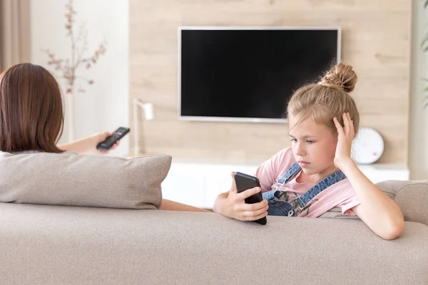 Mädchen sitzt auf der Couch und schaut etwas Interessantes im Handy, während Mutter fernsieht — Stockfoto