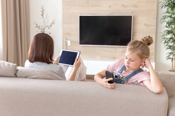 Chica sentada en el sofá y viendo algo interesante en el teléfono móvil mientras la madre viendo la televisión —  Fotos de Stock