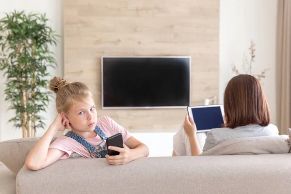 Mädchen sitzt auf der Couch und schaut etwas Interessantes im Handy, während Mutter fernsieht — Stockfoto