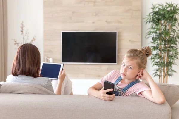 Chica sentada en el sofá y viendo algo interesante en el teléfono móvil mientras la madre viendo la televisión —  Fotos de Stock