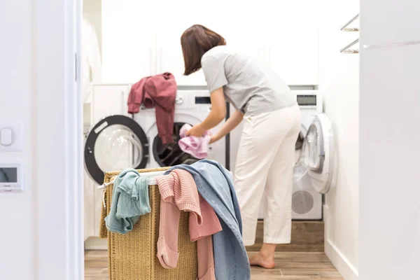 Mujer joven cargando lavadora y una cesta llena de ropa sucia en la sala de lavandería — Foto de Stock