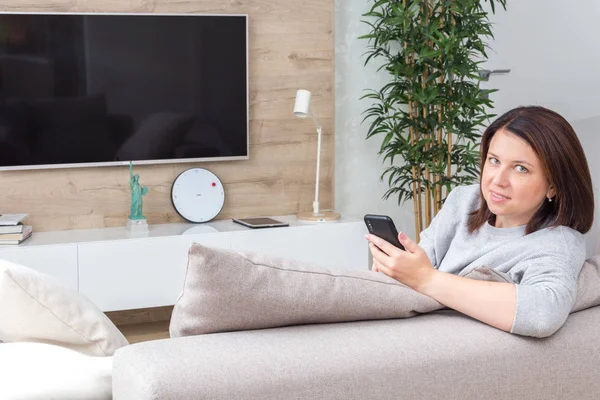 Молодая женщина смотрит на свой смартфон и улыбается сидя на диване — стоковое фото