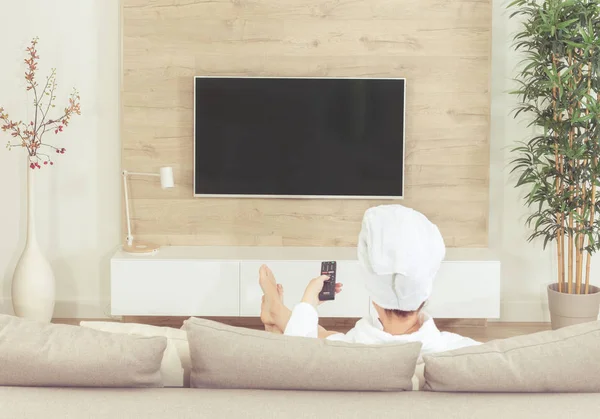 Mujer sentada en un sofá con toalla en la cabeza después de la ducha viendo la televisión —  Fotos de Stock