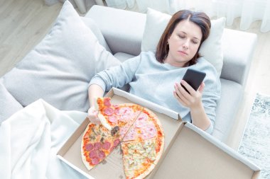Üzgün kadın pizza yiyor ve evde kanepede yatan telefon tutarak, mavi sesi