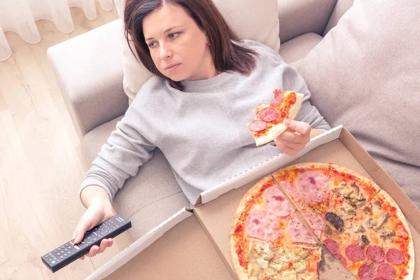Sad woman eating pizza and holding phone laying on sofa at home, warm yellow tone