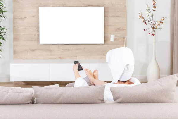 Duştan sonra başında havluyla kanepede oturan kadın televizyon izliyor. Tasarım için beyaz ekran kesiği — Stok fotoğraf