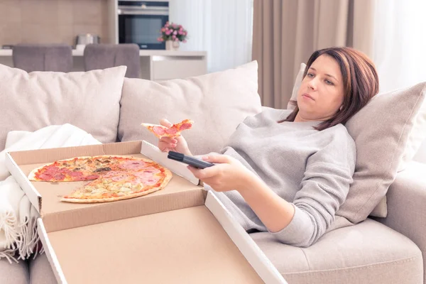 Sad woman eating pizza and holding phone laying on sofa at home, warm yellow tone