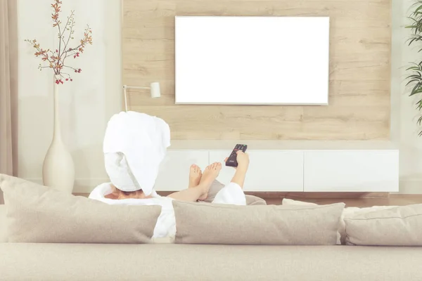 Mujer sentada en el sofá con toalla en la cabeza después de la ducha viendo la televisión. Pantalla blanca hecha para el diseño —  Fotos de Stock