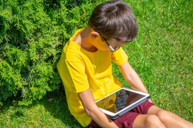 Tablet okuma elektronik kitap ile bir parkta çim üzerinde oturan çocuk