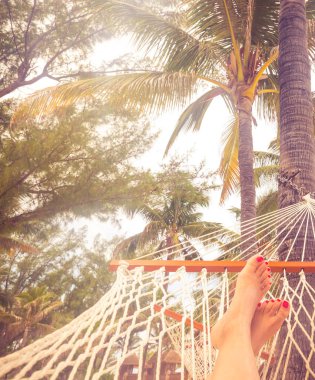 Deniz, palmiye ağaçları ve gün batımı bir arka plan üzerinde bir hamak kadın bacakları. Turuncu renk tonu ile tatil konsepti