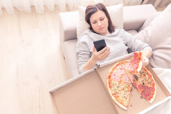 Депрессивная женщина ест пиццу и держит телефон, лежащий на диване дома, теплый желтый тон — стоковое фото