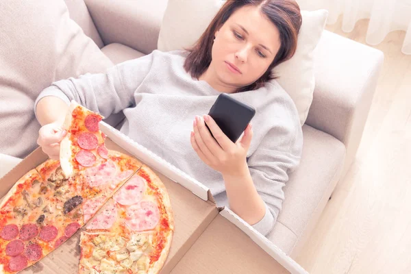 Depressed woman eating pizza and holding phone laying on sofa at home, warm yellow tone