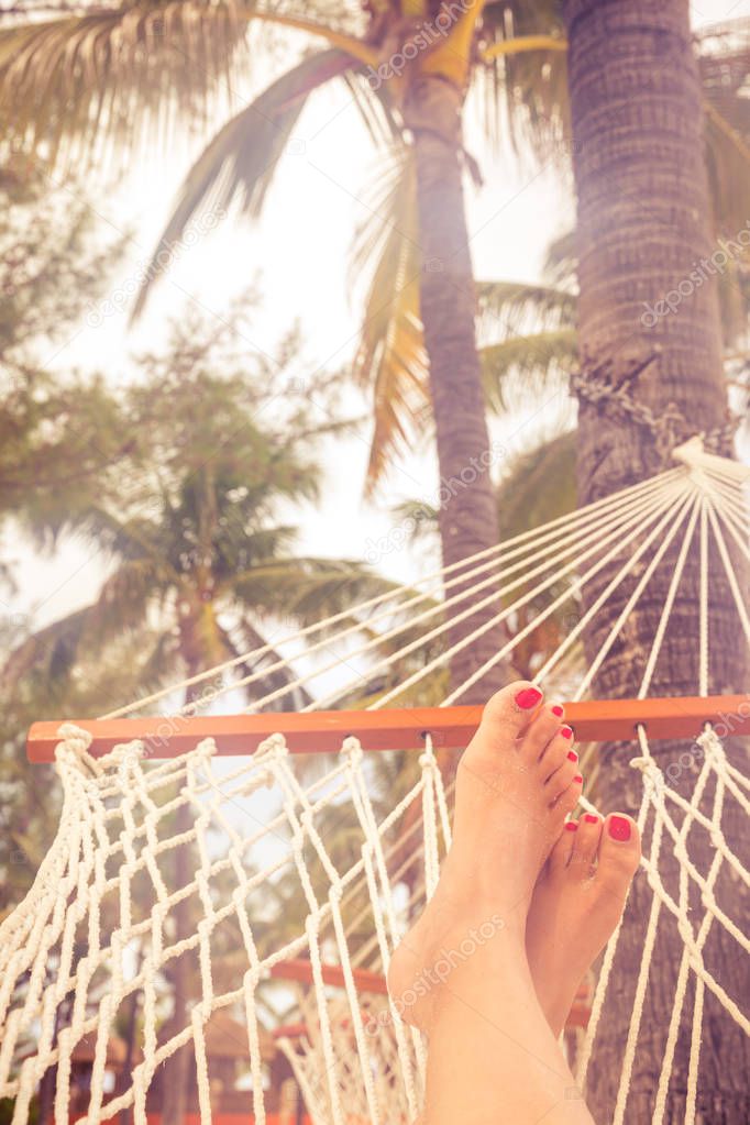 Female legs in a hammock on a background of the sea, palm trees and sunset. Vacation concept with orange color tone