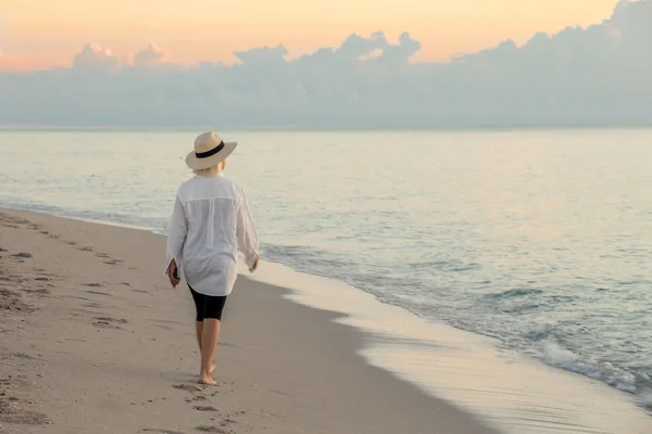 Mujer sosteniendo el teléfono celular y usando sombrero de paja caminando en una playa al atardecer —  Fotos de Stock