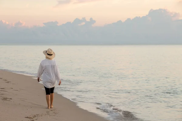 Mujer caminando a lo largo de la playa al atardecer sosteniendo teléfono celular y usando sombrero de paja —  Fotos de Stock