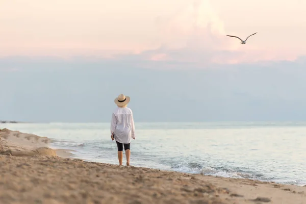 Frau, die bei Sonnenuntergang mit Handy am Strand entlang läuft und Strohhut trägt und träumt — Stockfoto