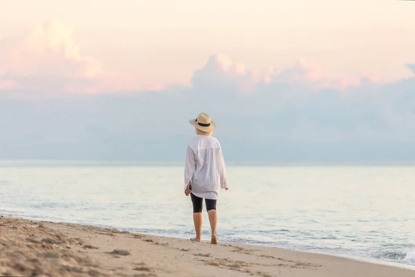 Frau mit weißem Hemd und Strohhut spaziert bei Sonnenuntergang am Strand — Stockfoto