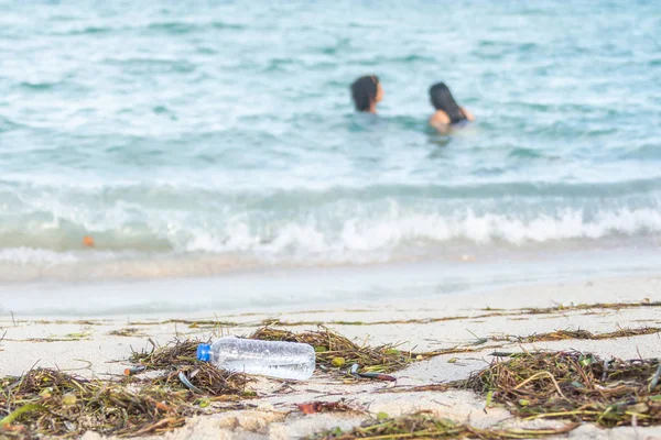 Close up image of empty plastic water bottle on dirty beach filled with seaweed, garbage and waste on dirty sandy beach with people in sea on background — Stock Photo, Image